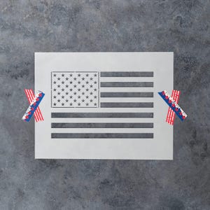 May include: A rectangular white stencil featuring a cutout design of the American flag, including the stars in the canton and horizontal stripes. The stencil is secured on both sides by two pieces of decorative tape with red, white, and blue patterns.