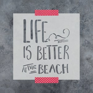 May include: A white stencil with the text "Life is better at the beach" in black lettering. The stencil is taped to a gray textured surface.