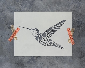 May include: A rectangular stencil featuring a cutout of a hummingbird in flight with intricate wing and body patterns. The stencil is secured on the left and right sides with two pieces of orange and gold patterned adhesive tape.