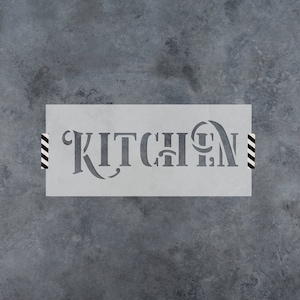 May include: A gray stencil with the word "KITCHEN" cut out in a decorative font. The stencil is rectangular and taped to a textured gray surface with black and white striped tape.