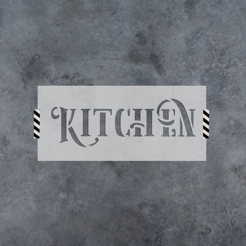 May include: A gray stencil with the word "KITCHEN" cut out in a decorative font. The stencil is rectangular and taped to a textured gray surface with black and white striped tape.