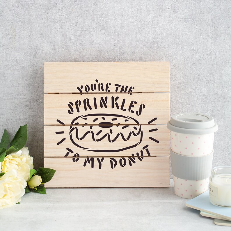 Op de afbeelding: Een houten bord met de tekst "You're the sprinkles to my donut" met een donut-illustratie. Het bord staat op een grijze achtergrond met een grijs-witte stippenbeker en een witte kaars.
