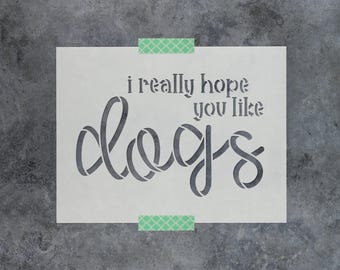 May include: A rectangular stencil featuring the text i really hope you like dogs in a combination of block and cursive lettering. The stencil is white with cut-out letters and is secured at the top and bottom with strips of green patterned tape.