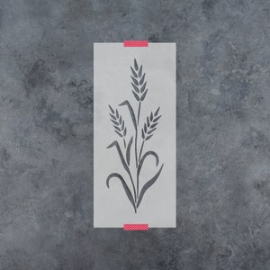 May include: A white stencil with a design of three wheat stalks. The stencil is taped to a gray textured surface.