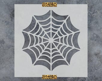 May include: A square, light gray stencil featuring a centered, die-cut spider web design with radiating lines and concentric arcs. The stencil is secured at the top and bottom edges with two strips of adhesive tape displaying a brown leopard print.