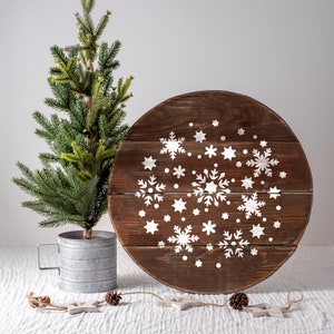 K&ouml;nnte beinhalten: Ein rundes Holzschild mit einem wei&szlig;en Schneeflockenmuster. Das Schild ist braun und hat einen rustikalen Look. Im Vordergrund steht ein kleiner gr&uuml;ner Weihnachtsbaum.