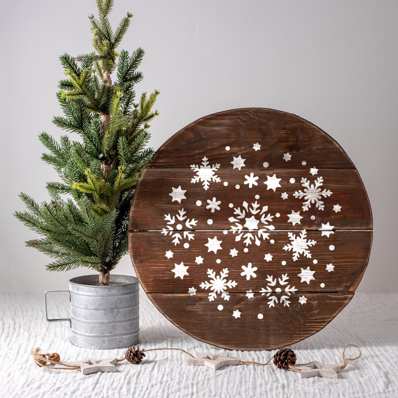K&ouml;nnte beinhalten: Ein rundes Holzschild mit einem wei&szlig;en Schneeflockenmuster. Das Schild ist braun und hat einen rustikalen Look. Im Vordergrund steht ein kleiner gr&uuml;ner Weihnachtsbaum.