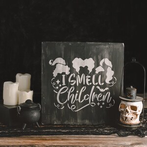 May include: A black wooden sign with a white stencil design featuring three witches and the text "I smell children". The sign is sitting on a wooden table with a black cauldron and a skull lantern.