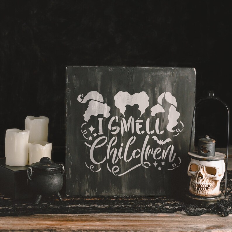 May include: A black wooden sign with a white stencil design featuring three witches and the text "I smell children". The sign is sitting on a wooden table with a black cauldron and a skull lantern.