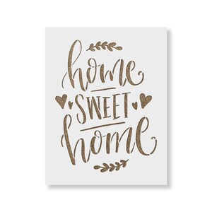May include: A white stencil with the text "home sweet home" in brown lettering. The text is in a cursive font and is surrounded by two small hearts and two sprigs of leaves.