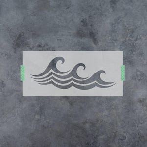 May include: A rectangular stencil featuring a stylized graphic of three ocean waves. The stencil is light gray with a cutout design of the waves and has small green patterned tape strips on the left and right edges to secure it in place.
