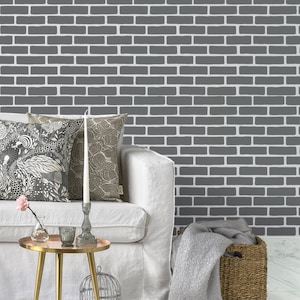 May include: A white linen sofa with two patterned throw pillows and a white candle on a silver candlestick. The sofa is in front of a gray brick wall. A gold side table with a round top and three legs is in front of the sofa. A gray throw blanket and a wicker basket are on the floor in front of the sofa.