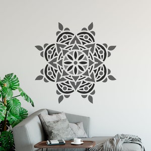 May include: A large, gray and black mandala wall decal. The symmetrical design features a central floral pattern surrounded by layered geometric shapes. The decal is mounted on a white wall above a gray sofa, adding a decorative element to the living space.