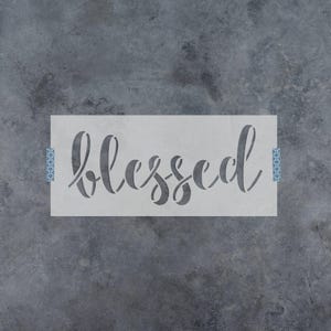 May include: A rectangular stencil featuring the word blessed in a cursive script font. The stencil is light gray and is secured on the left and right sides with small strips of blue and white patterned tape against a dark gray textured surface.