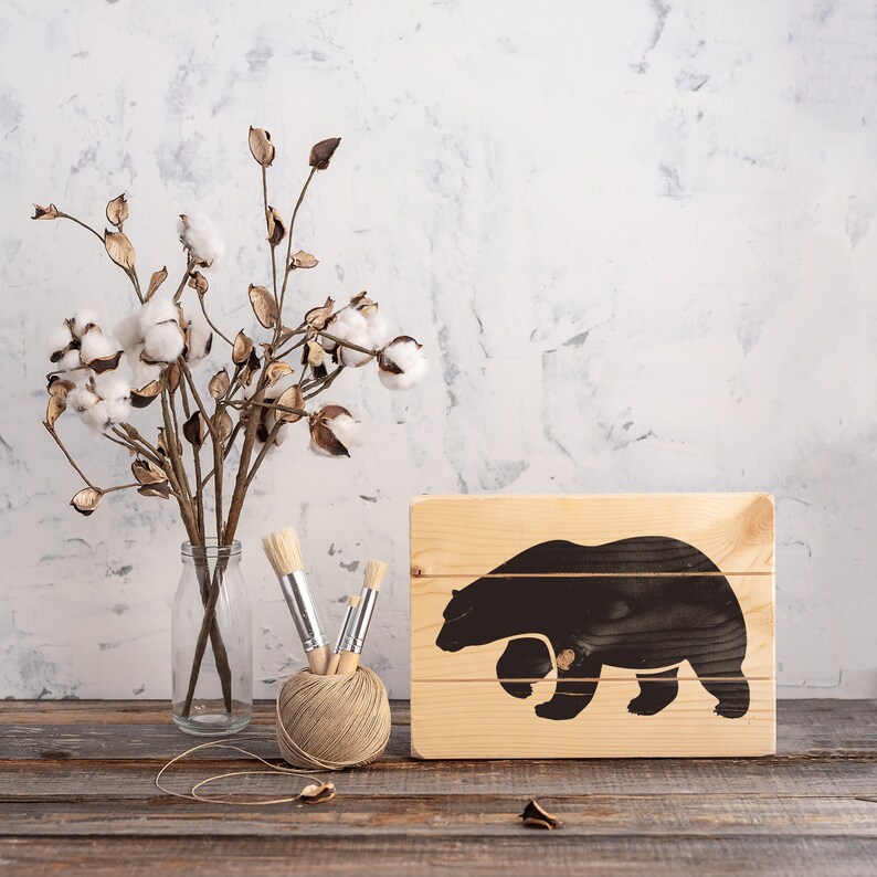 May include: A wooden sign featuring a black silhouette of a bear walking to the right. The sign is placed on a wooden table with a vase of dried cotton flowers and paintbrushes.