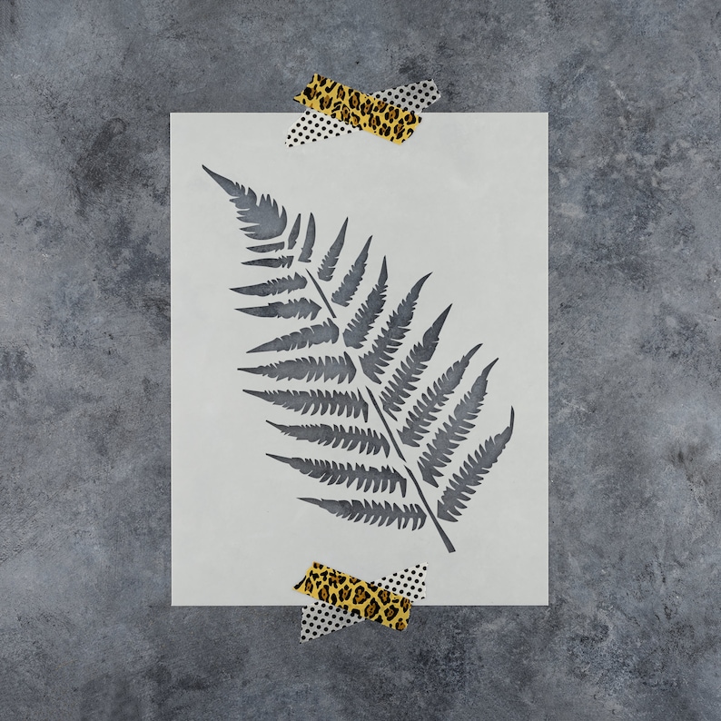 May include: A white stencil featuring a fern leaf design. The stencil is taped to a grey background.