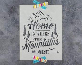 May include: A rectangular stencil featuring a mountain range graphic at the top and the text Home IS WHERE THE Mountains ARE in a mix of serif and script fonts. Pine trees flank the text, and two pieces of colorful striped tape secure it.