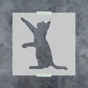 May include: A square stencil featuring a dark gray silhouette of a cat standing on its hind legs with one front paw raised upward. The stencil is made of a translucent material and is secured at the top and bottom edges with small pieces of tape.