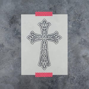 May include: A rectangular stencil sheet featuring a central ornate cross design with intricate filigree cutouts. The stencil is held in place by two strips of pink and white polka dot tape at the top and bottom edges of the white rectangular sheet.