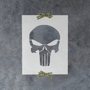 May include: A white rectangular stencil featuring a large, centered skull design with hollowed-out eye sockets and a vertical-striped jaw. The stencil is secured at the top and bottom with two pieces of patterned, olive-green camouflage tape.