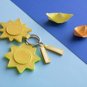 Puede incluir: Llaveros amarillos con forma de sol con un borde verde neón y el texto "ASILEATHER" en la etiqueta. Los llaveros están en un anillo plateado. También hay dos barcos de papel origami en la imagen, uno amarillo y otro naranja.