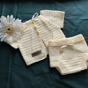 May include: Cream-colored, crocheted baby outfit set, including a short-sleeved cardigan with three yellow buttons and matching shorts. The set is adorned with cream-colored ribbons and a small tag that reads "Sockenreich". A white gerbera daisy rests near the outfit.