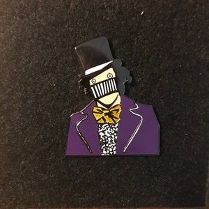 May include: A purple and black enamel pin featuring a cartoon character wearing a top hat, a bow tie, and a black and white striped mask. The character has a white face and black eyes.