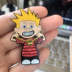 May include: A cartoon-style enamel pin featuring a character with spiky, glittery gold hair and a red and black striped shirt. The character is wearing a gold chain and has an angry expression. The pin has white feet.