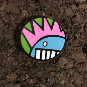 May include: A round, colorful enamel pin featuring a cartoon creature with a pink mohawk, green and white stripes, a blue body, and a white mouth with black teeth.