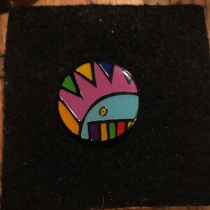May include: A colorful enamel pin featuring a cartoon creature with a pink mohawk, blue body, and a rainbow mouth.
