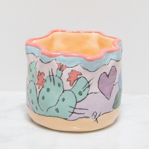 May include: A small, hand-painted ceramic planter with a scalloped rim. The planter features a whimsical design of a cactus with pink flowers, a purple heart, and a blue flower. The planter is painted in pastel colours on a light pink background.