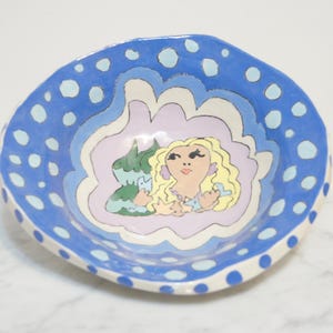 May include: A hand-painted ceramic bowl with an irregular, wavy rim. The exterior and rim feature a bright blue glaze with light blue polka dots. The interior center displays a painted illustration of a blonde figure with green accents.
