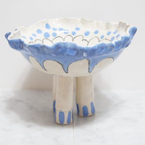 Pode incluir: Uma tigela de cerâmica com base branca e detalhes azuis. A borda é ondulada com uma borda azul e pontos azuis. Ela é suportada por três pernas curtas e cilíndricas, também com detalhes azuis. Aproximadamente 25 cm de diâmetro.