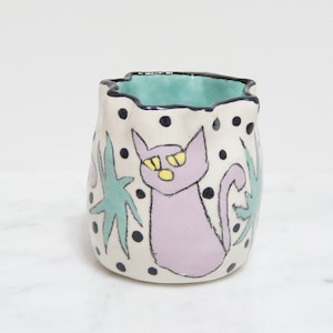 May include: A white ceramic cup with a green and black rim. The cup has a hand-painted design of a purple cat with yellow eyes and green leaves on a white background with black polka dots.