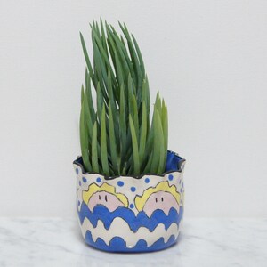 Puede incluir: Una maceta de cerámica pintada a mano con un diseño azul, blanco y amarillo con dos caras estilizadas. La maceta tiene un borde festoneado y está llena de una planta suculenta verde.