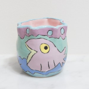 May include: A small, hand-painted ceramic planter with a scalloped rim. The planter is decorated with a pink fish with a yellow eye and a blue, green, and pink background.