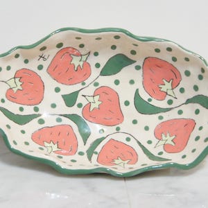 May include: Oval ceramic serving dish with a scalloped edge. The dish is cream-colored with a pattern of red strawberries, green leaves, and green polka dots. The rim is painted green.