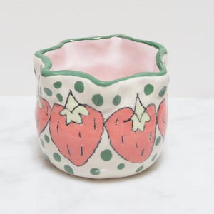 May include: A ceramic container with a wavy rim, featuring a hand-painted strawberry design. The strawberries are red with green tops, set against a cream background with green polka dots. The interior is pink, and the rim is outlined in green.