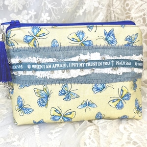 May include: A rectangular, cream-colored pouch with a blue zipper and tassel. The pouch is decorated with blue and yellow butterflies. A blue denim band with white lace and the text "WHEN I AM AFRAID, I PUT MY TRUST IN YOU PSALM 56:3" is sewn across the middle.