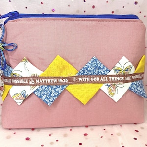 May include: A pink fabric pouch with a blue zipper and a decorative zig-zag pattern. The pattern includes yellow, blue floral, and butterfly-printed fabric. A ribbon with the text "WITH GOD ALL THINGS ARE POSSIBLE" is across the center.