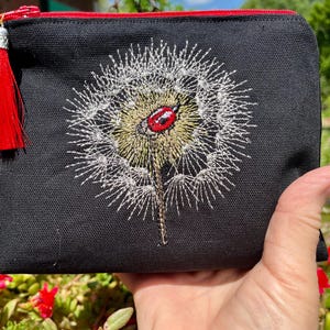 May include: Black zippered pouch with a red tassel. The pouch features an embroidered dandelion with a ladybug. The zipper is red, and the top edge of the pouch is trimmed in red.