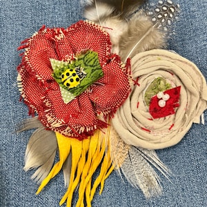 May include: A handmade fabric flower arrangement with a red burlap flower, a cream-colored flower, and yellow accents. The red flower features a green leaf with a yellow ladybug. Feathers in shades of gray and white add texture.