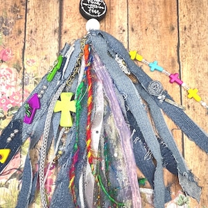 May include: A decorative tassel featuring frayed denim strips, colorful ribbons, and metallic chains. It includes a black circular bead with the text Faith Over Fear, a silver lobster clasp, and various charms including crosses in yellow, pink, and blue.