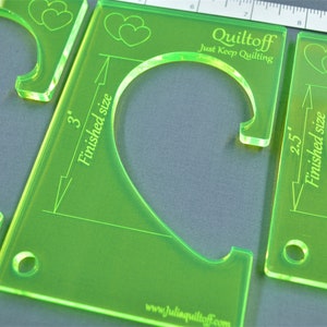 May include: Two green acrylic quilting templates with heart-shaped cutouts. The larger template is 3 inches and the smaller template is 2.5 inches. Both templates have the text "Quiltoff Just Keep Quilting" and a website address.