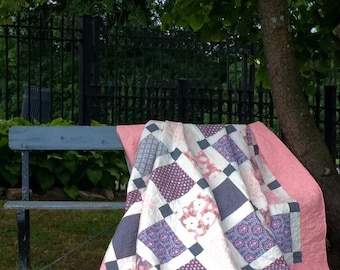 May include: A handmade quilt draped over a bench in a garden setting. The quilt features a patchwork design with squares of white, purple, and gray fabrics. The quilt is bordered with a solid pink fabric. The bench is painted blue.