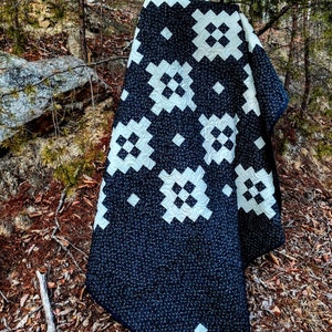 May include: A black and white quilt with a geometric pattern. The quilt is folded and draped over a rock in a wooded area.