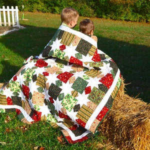 May include: Two children are sitting on a bale of hay, wrapped in a colourful patchwork quilt with a white background and green, red, and brown squares. The quilt has a star pattern.