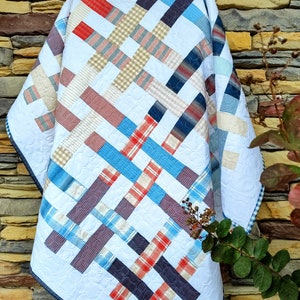 May include: A white quilt with a patchwork design featuring blue, red, and tan plaid fabric. The quilt is folded and leaning against a stone wall.
