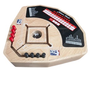 May include: A wooden baseball dice game board featuring a central dice cup, four red pegs, and four black pegs. The board includes a scoreboard labeled Johnson Creek MAPACHES, DCL logos, and printed game instructions for various baseball outcomes.