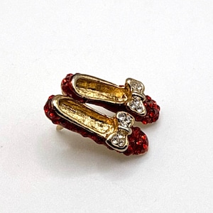 Peut inclure: Une paire de chaussures miniatures rouges recouvertes de strass, avec des intérieurs et des nœuds dorés, rappelant les chaussons rouges de Dorothy. Un support de broche doré est également visible.